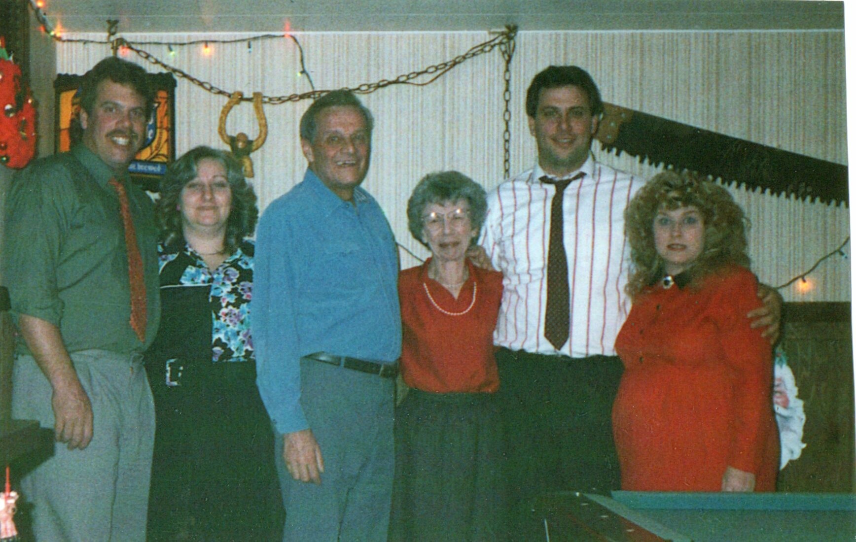 Six adults posing together at a holiday party, standing indoors with Christmas lights in the background and a festive atmosphere.