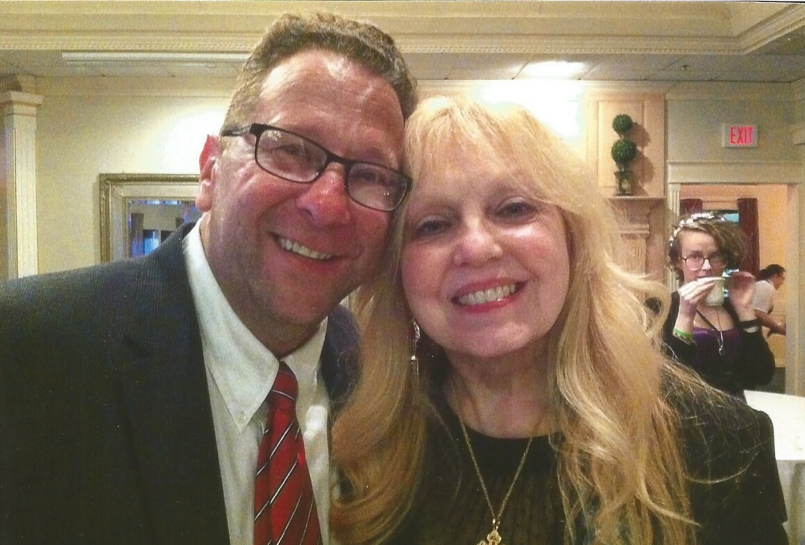 A man in a suit and red tie stands beside a smiling woman with long blonde hair at an indoor event.