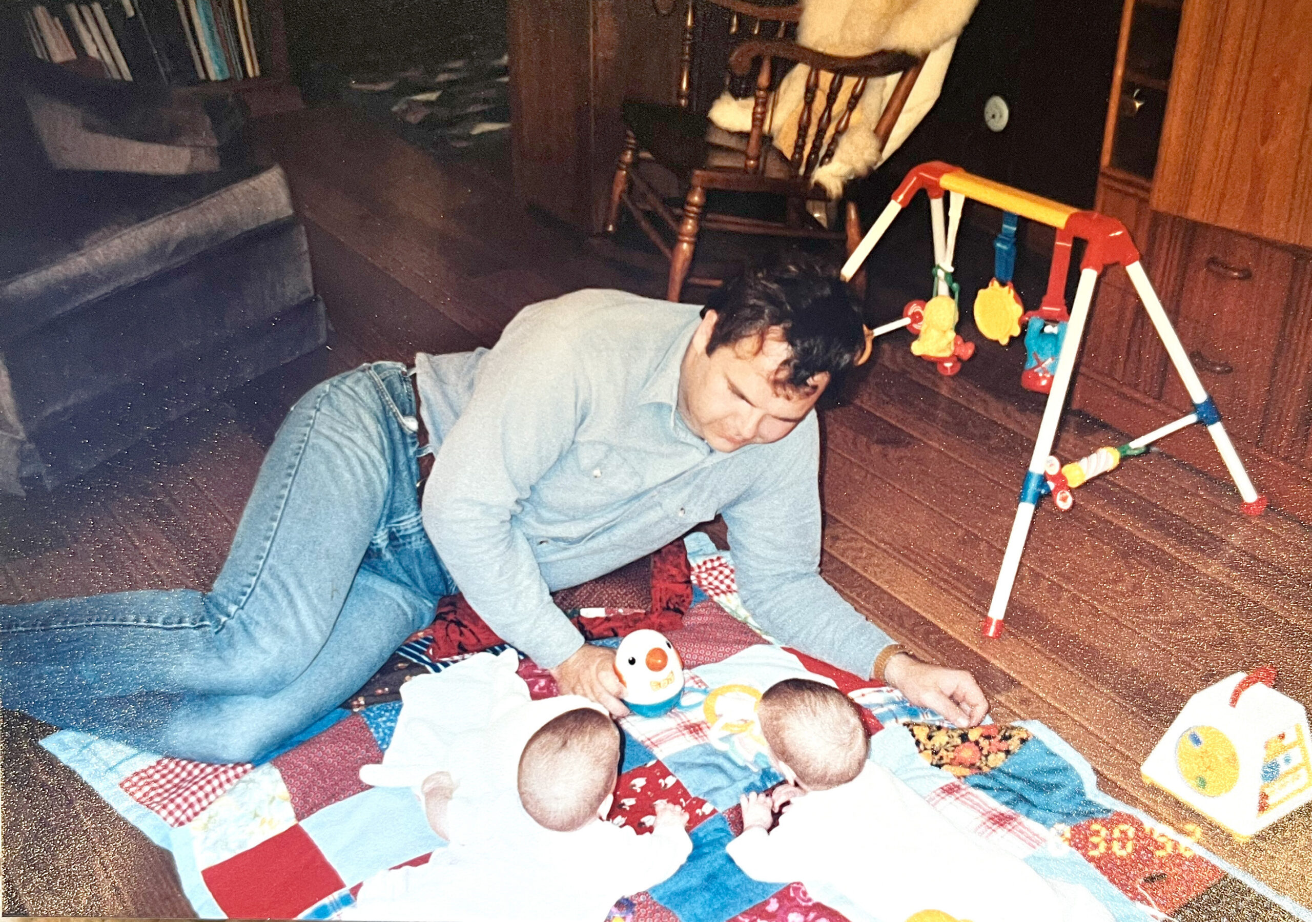 Child lying on a patterned play mat on the floor, reaching for small toys nearby with a colorful baby activity gym in the background.