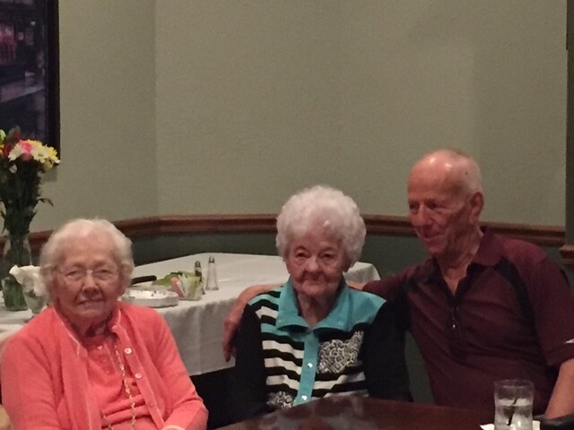 Three elderly adults sit together at a dining table in a restaurant, smiling for the camera. (woman in coral cardigan left, woman with teal/black striped jacket center, man in maroon shirt right)