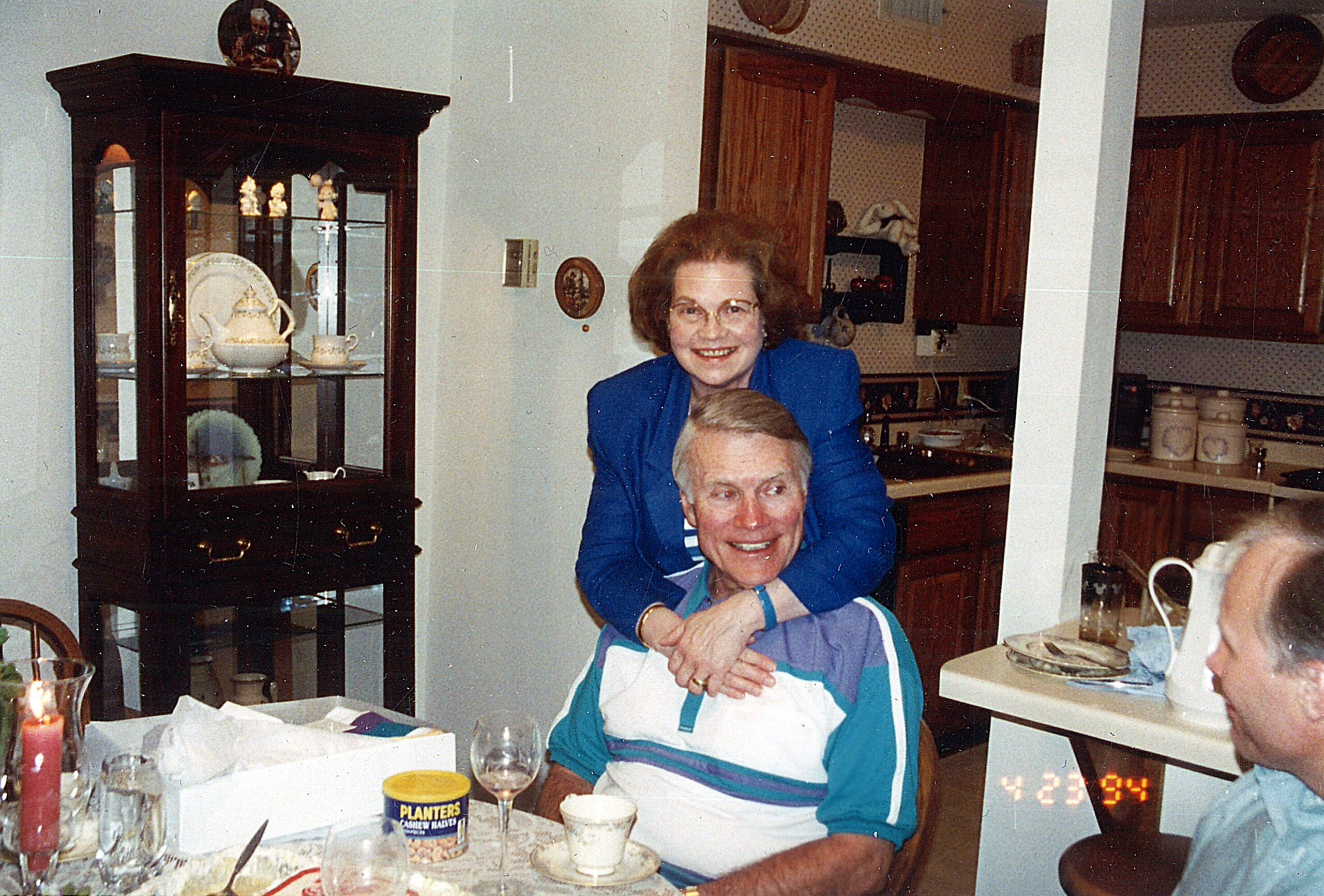 Group of people at a dining table in a home kitchen, smiling and posing for a photo; woman in blue behind man in foreground, date stamp 1994 visible on right.