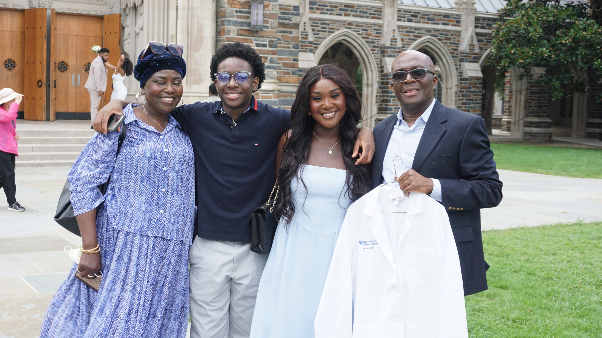 Group of four adults posing together outdoors on a university campus, one in a light blue graduation gown, others in smart casual clothing, all smiling at the camera.