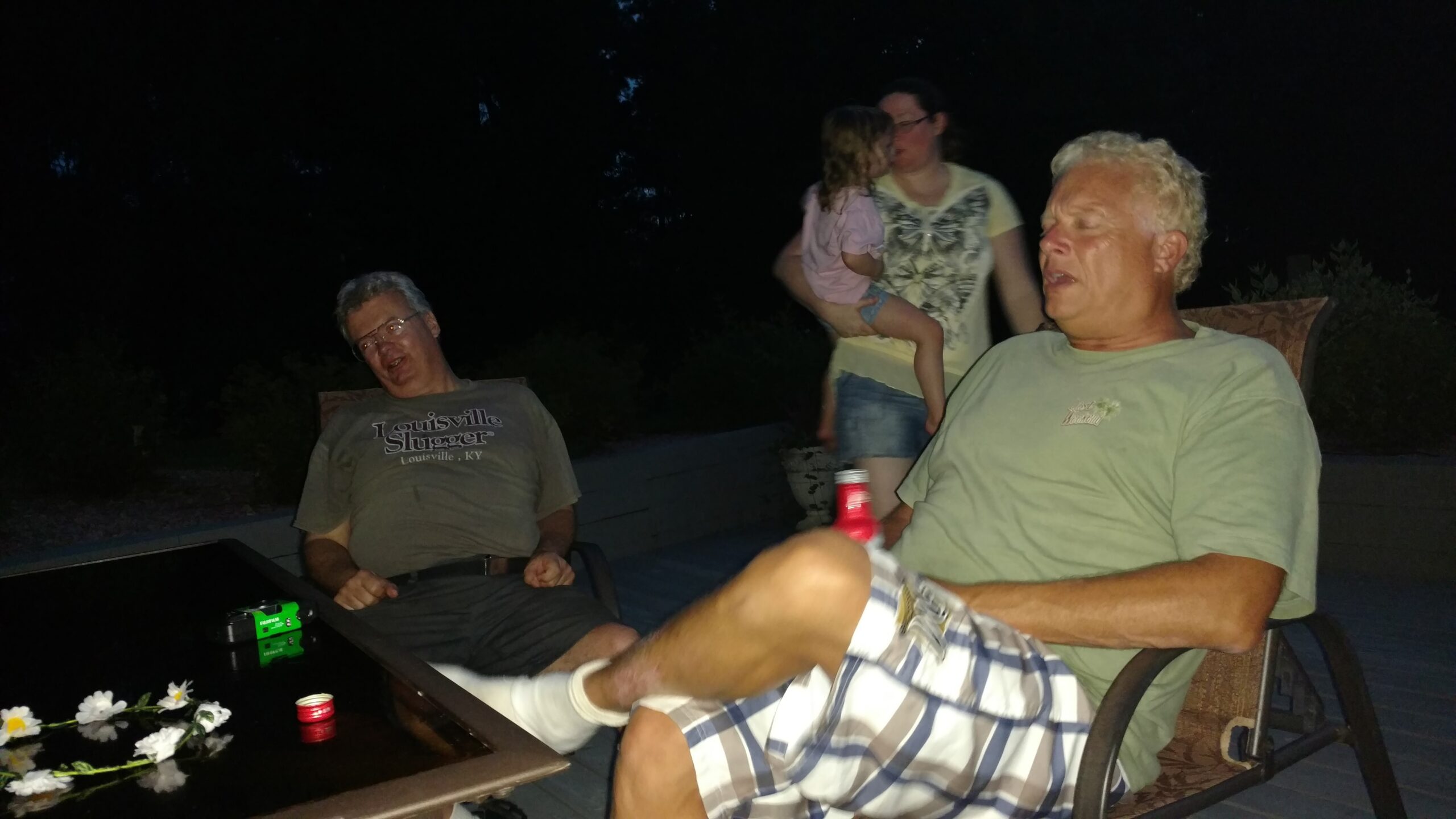 Group of adults outside at night; man with light hair in the foreground, another man laughing on the left, a woman holding a child in the background.