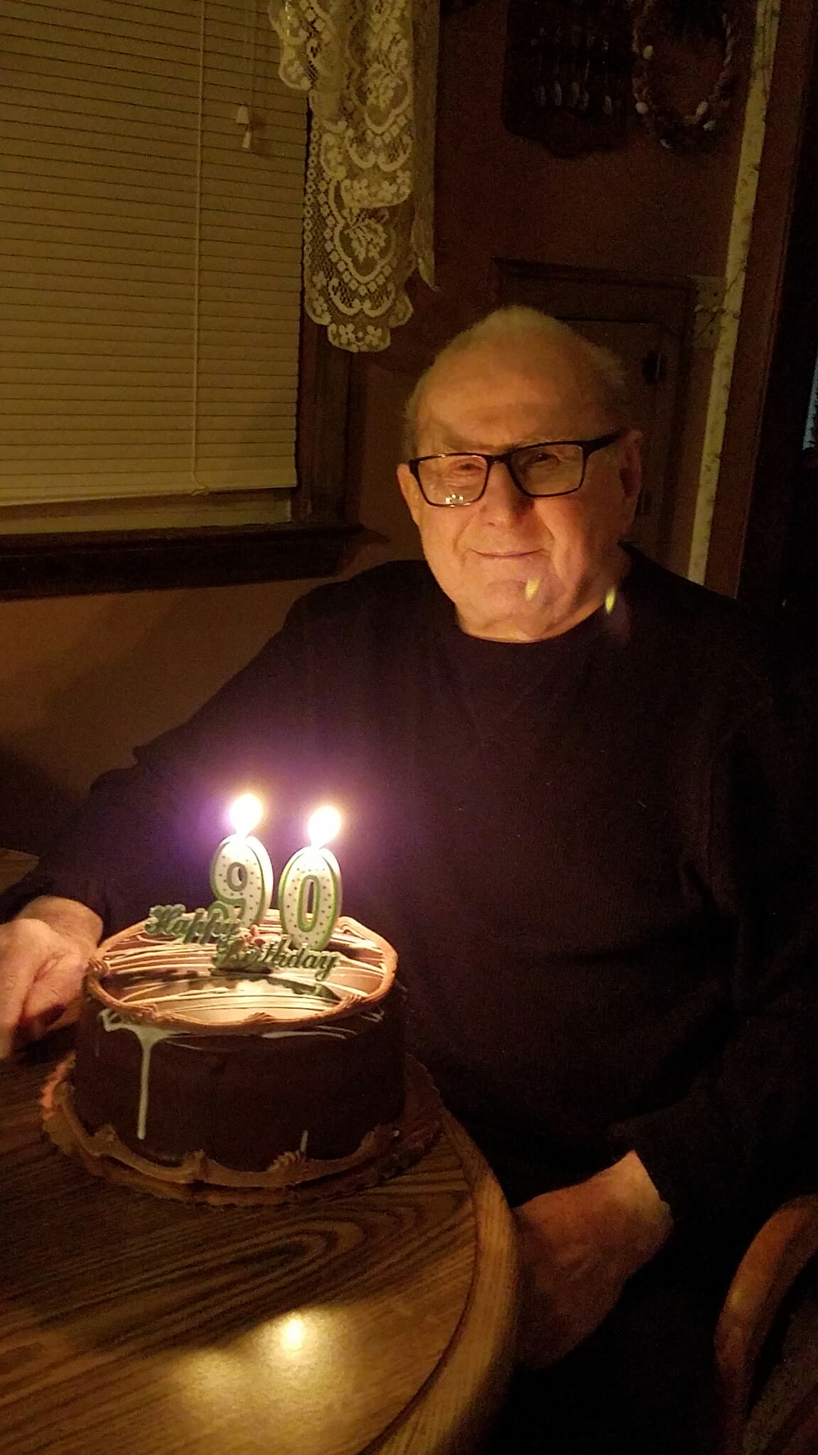 Smiling elderly man with glasses at a 90th birthday party, candles on a chocolate cake in front of him