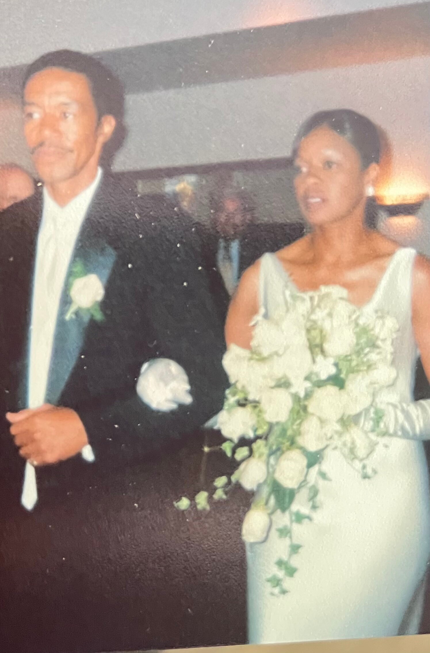 Older man in a black suit with a white boutonniere standing beside a woman in a pale dress holding a large white floral bouquet at a formal event.