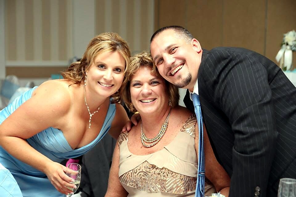 Three smiling people posing for a photo at a formal event: two women and a man leaning in together, with the woman on the left in a blue dress holding a glass.]