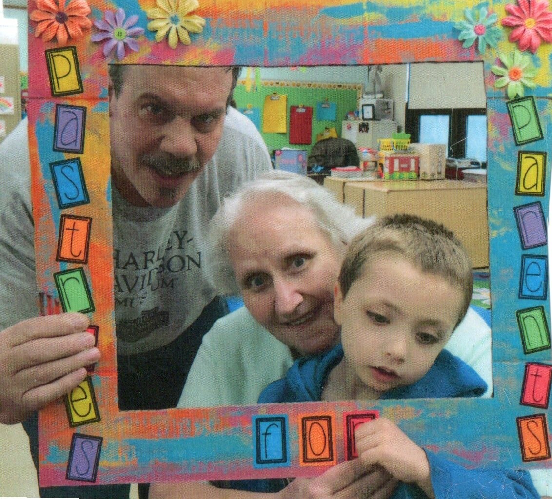 Family of three posing behind a colorful handmade photo frame decorated with flowers and letter tiles in a classroom.