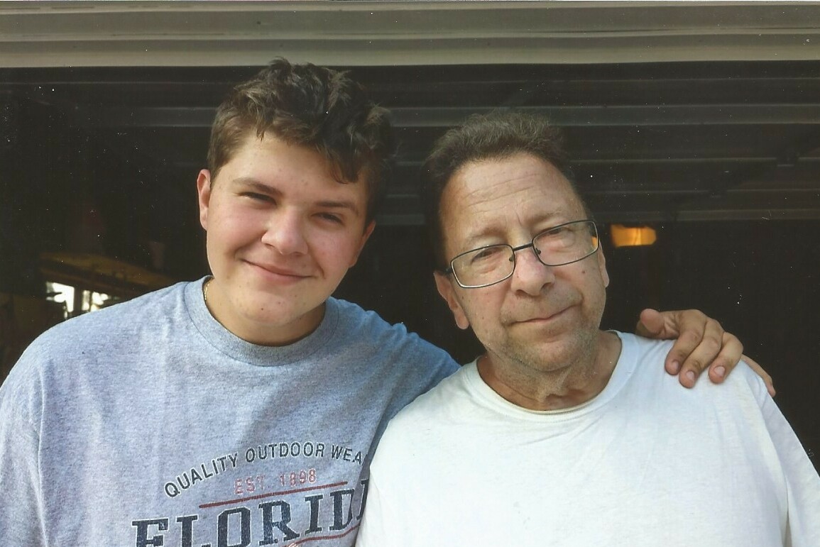 Two men posing together, one younger with short hair in a gray shirt and one older man with glasses in a white shirt, arm around the other in a garage doorway.