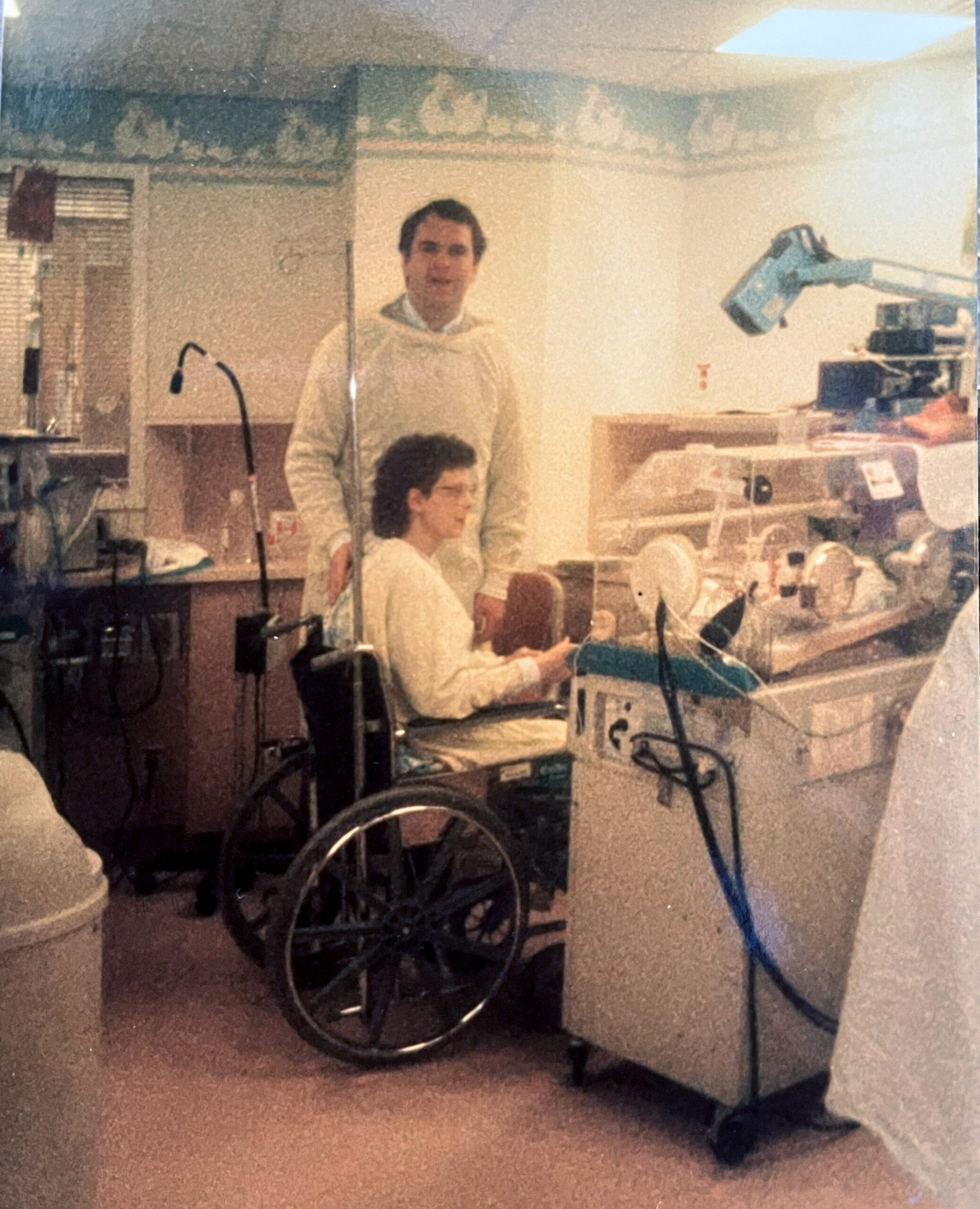 Two people in a clinical or lab setting, one seated and the other standing, with lab equipment including a blue robotic arm on the right and various instruments in the background.