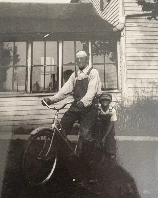 An elderly man in overalls stands beside a bicycle in front of a wooden house, with a young boy beside him.