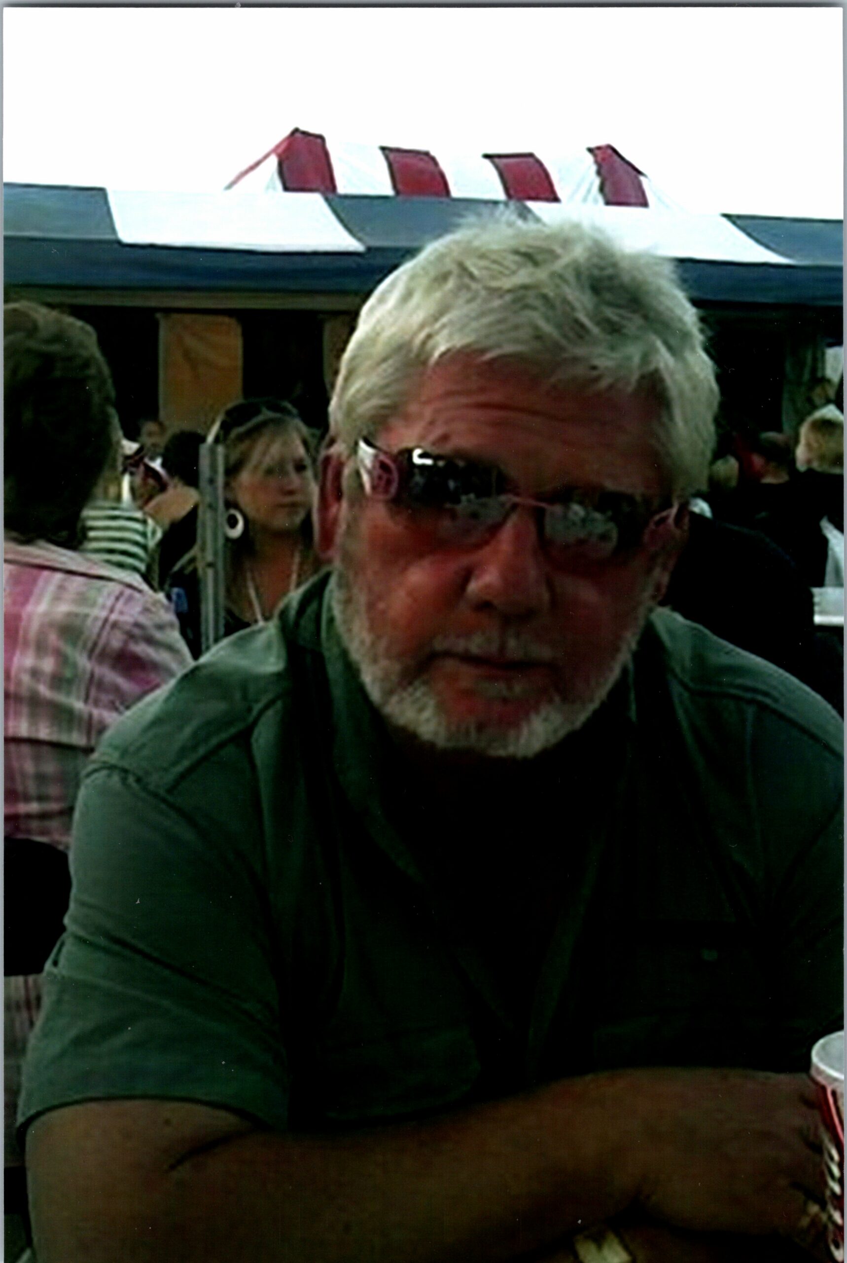 Close-up of an older man with gray hair and beard sitting at a table at an outdoor crowd event, with a white tent and red pennants in the background.