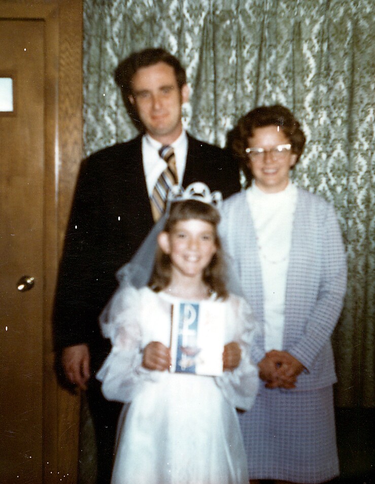 Young girl in a white dress and veil stands in front of two adults, holding a booklet and posing for a family photo in a room with patterned curtains behind them.
