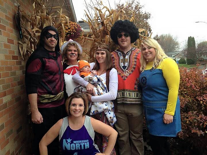 Group of six adults in colorful costumes posing outside with a baby in a carrier, fall corn stalks in the background.