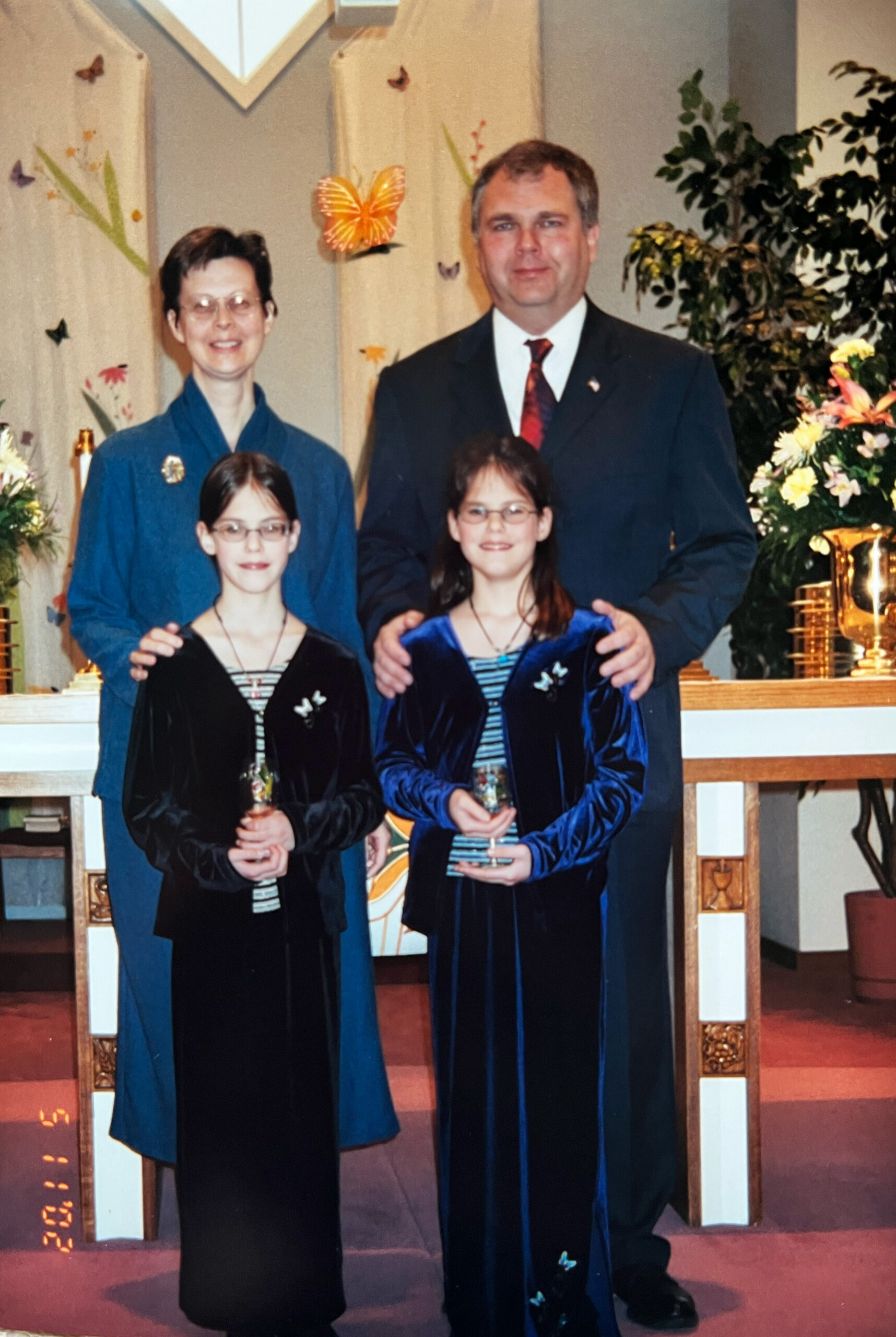 Family of four posing for a photo at an awards ceremony; two young girls in velvet dresses hold trophies in front, flanked by their parents.
