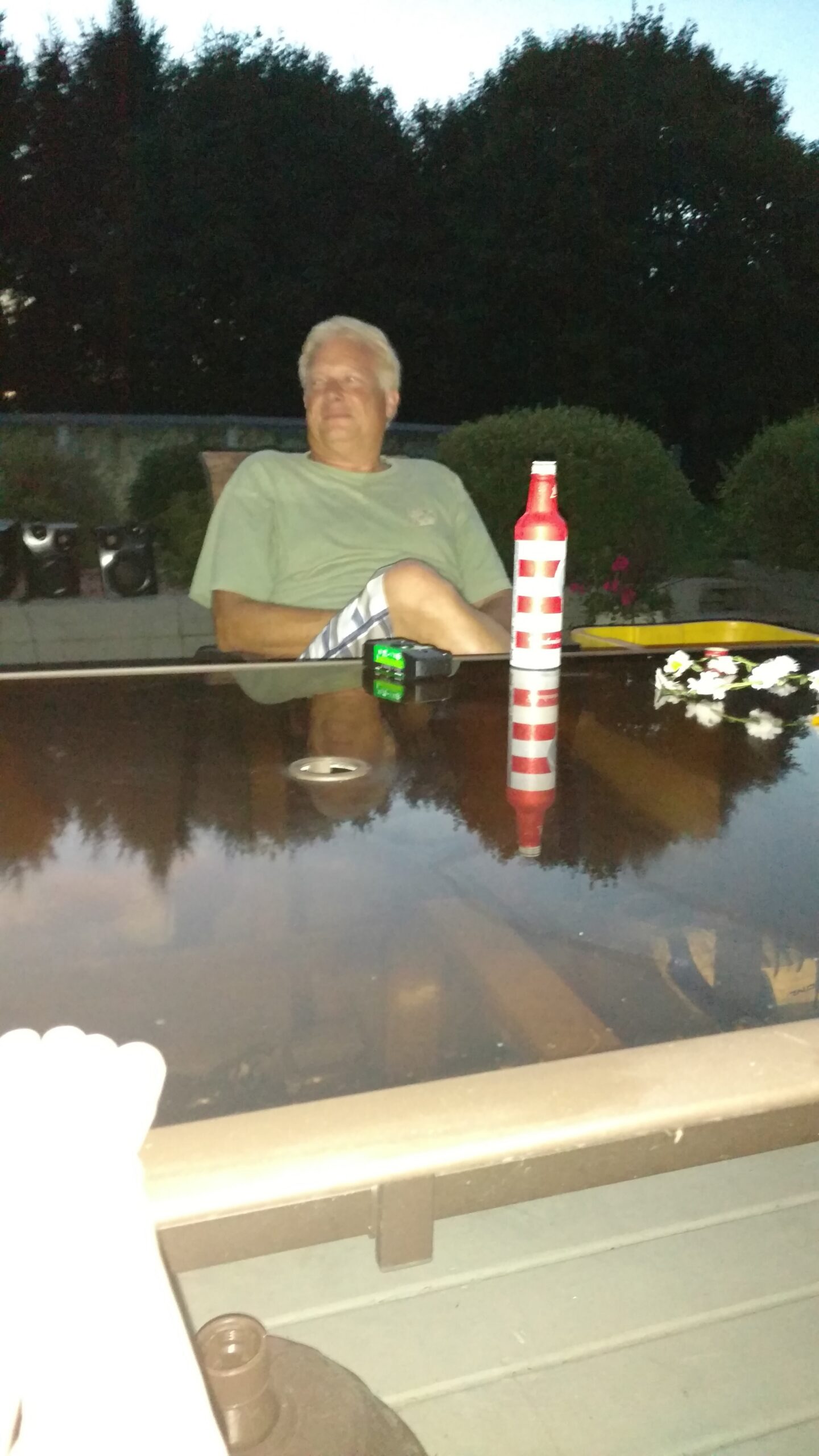 Older man in a green shirt sitting outside at a patio table, with a red-and-white striped bottle on the glass tabletop.