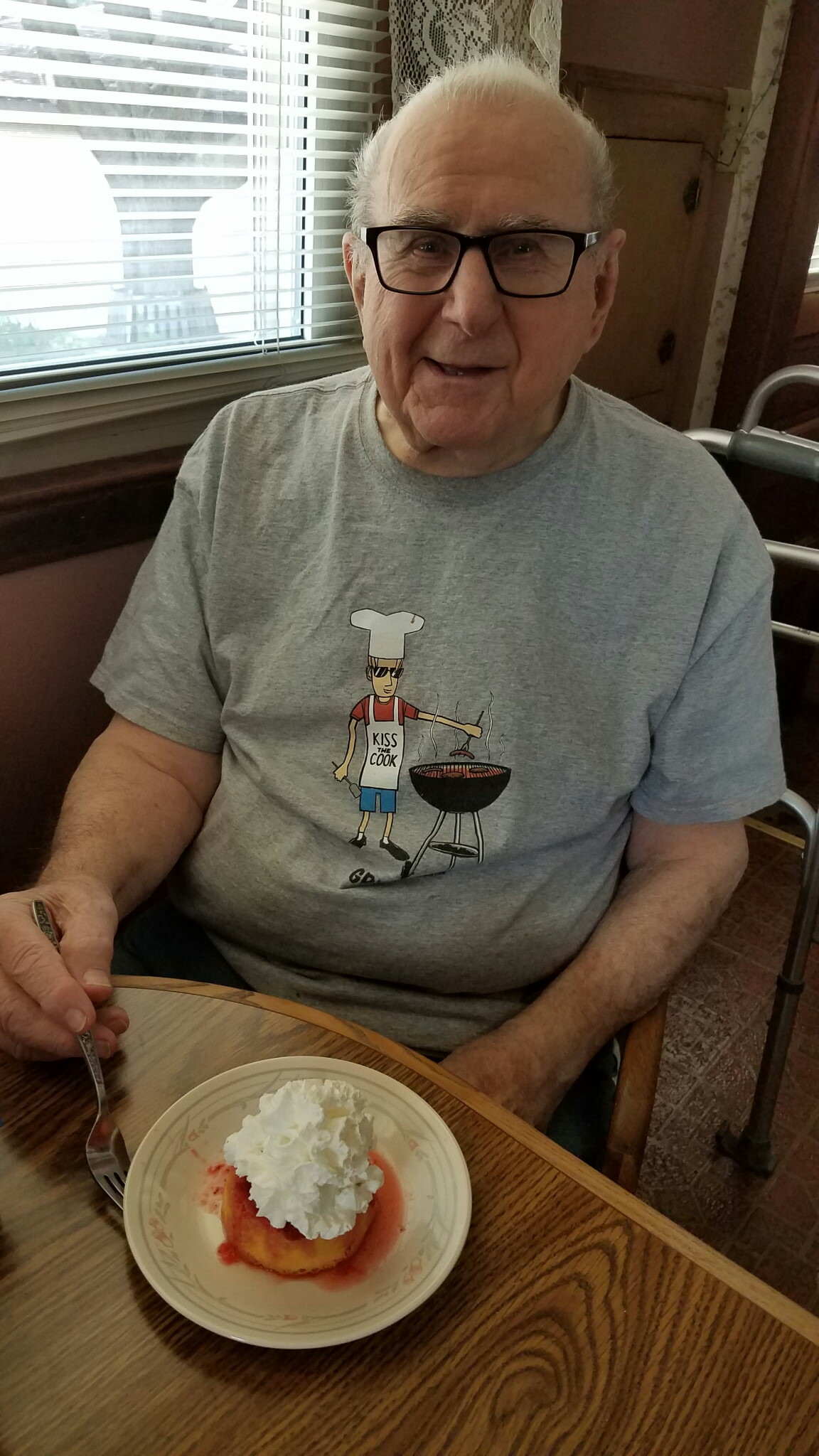 Smiling elderly man wearing glasses sits at a table, wearing a 'Kiss the Cook' chef T‑shirt, with a dessert plate in front of him.