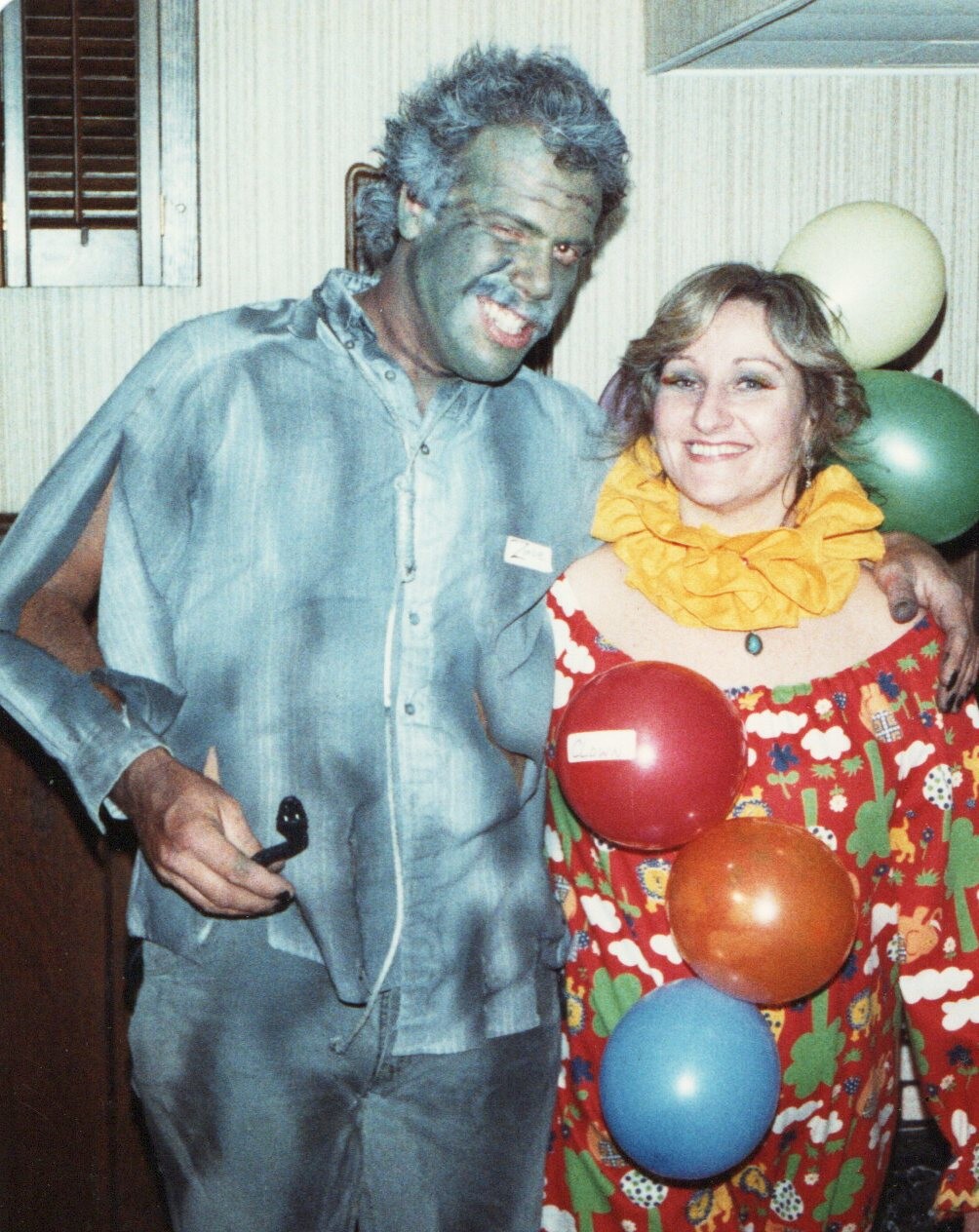 Two people at a party in costumes: a man with blue face makeup and gray hair in a light blue shirt, and a smiling woman in a colorful clown outfit with a yellow ruffled collar, with balloons in the background.