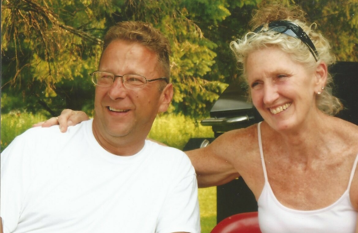 Man wearing glasses and a white shirt smiles with a woman in a white tank top outdoors, with greenery in the background.