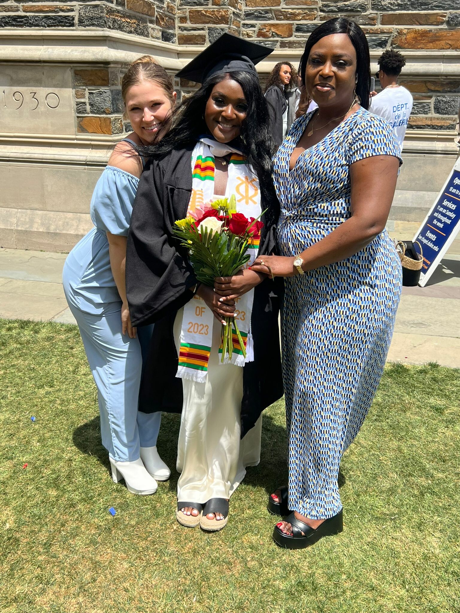 Graduating student in cap and gown holding a bouquet, flanked by two friends outdoors in front of a stone building.