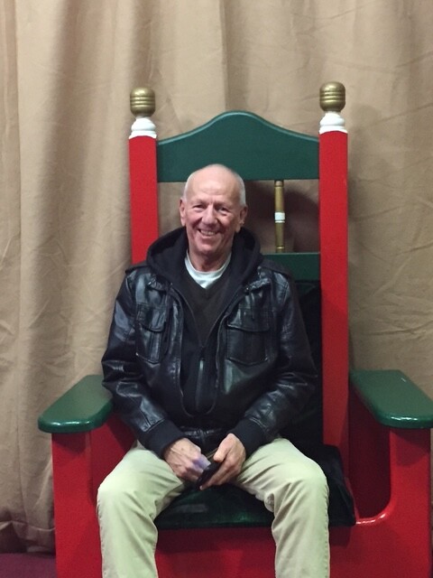 Smiling older man sitting on a large red and green throne-like chair indoors, wearing a black leather jacket and beige pants.