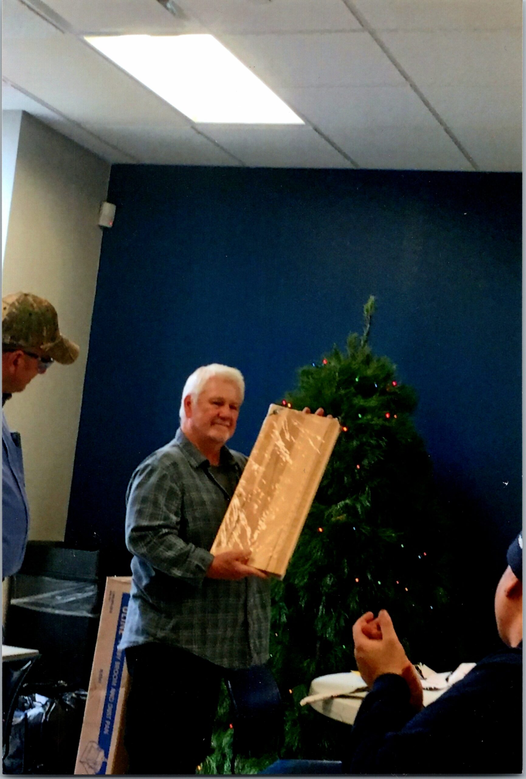 Older man in a plaid shirt smiles while receiving a long wrapped gift, with a Christmas tree in the background.