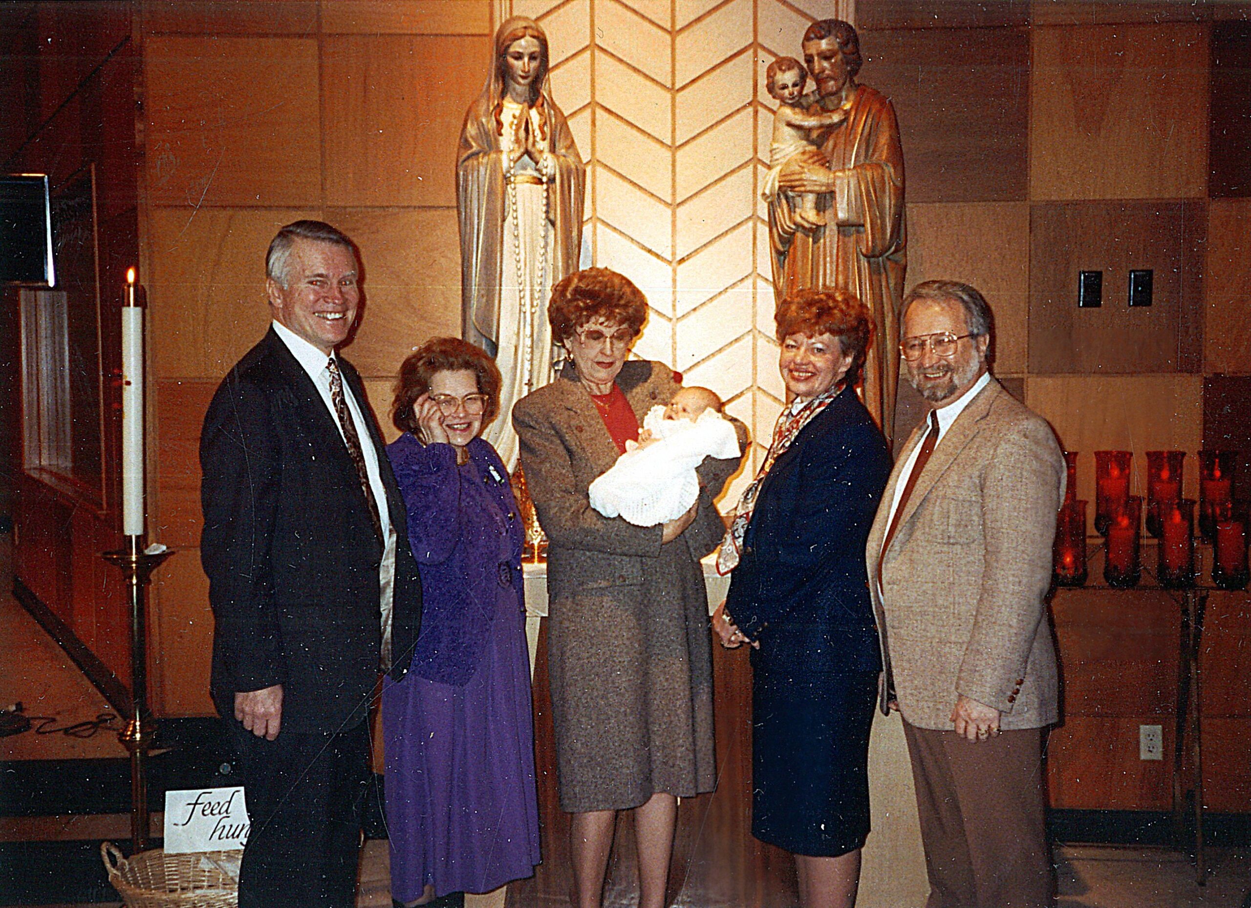 A family of five adults and a baby pose in a church in front of statues of Mary and Joseph with baby Jesus.