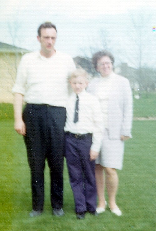 Family posing outdoors on a grassy lawn: tall man in white shirt and black pants, a boy in a white shirt and tie, and a woman in a light dress stand together with houses in the background.