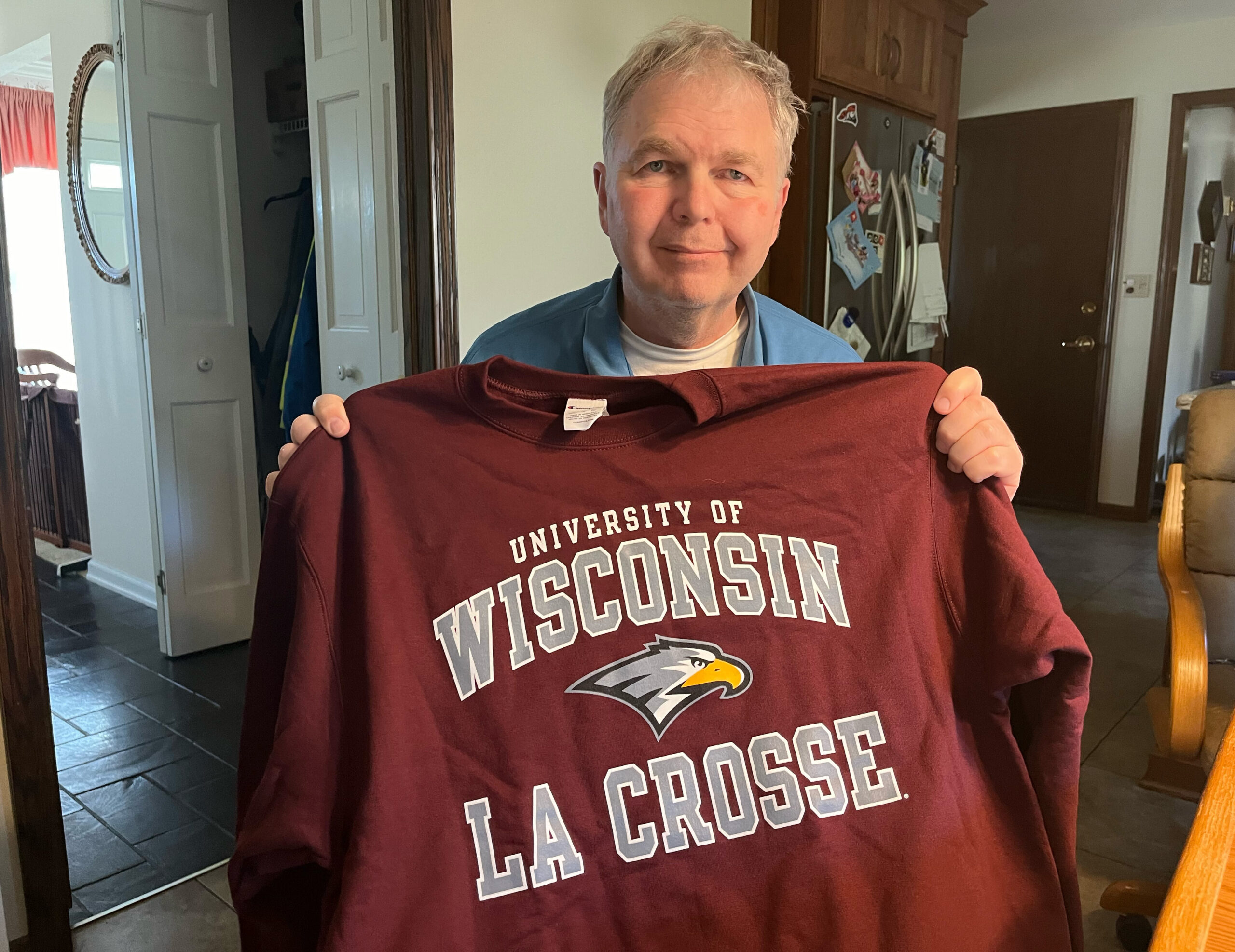 Smiling man holding up a maroon University of Wisconsin–La Crosse T‑shirt indoors.