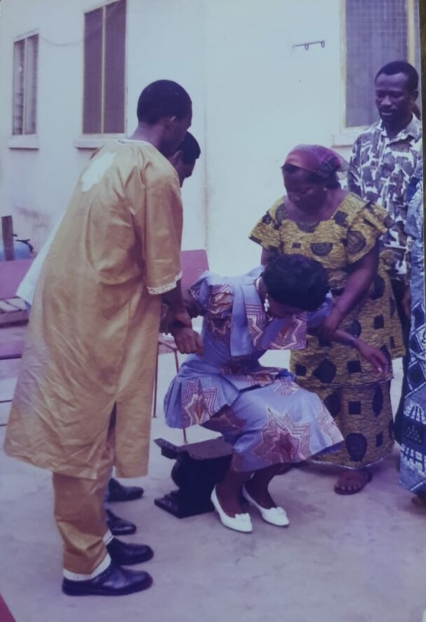 A group of people assist a seated woman in a purple patterned dress outdoors; a man in yellow robes and a woman in a yellow dress help her stand from a low chair.