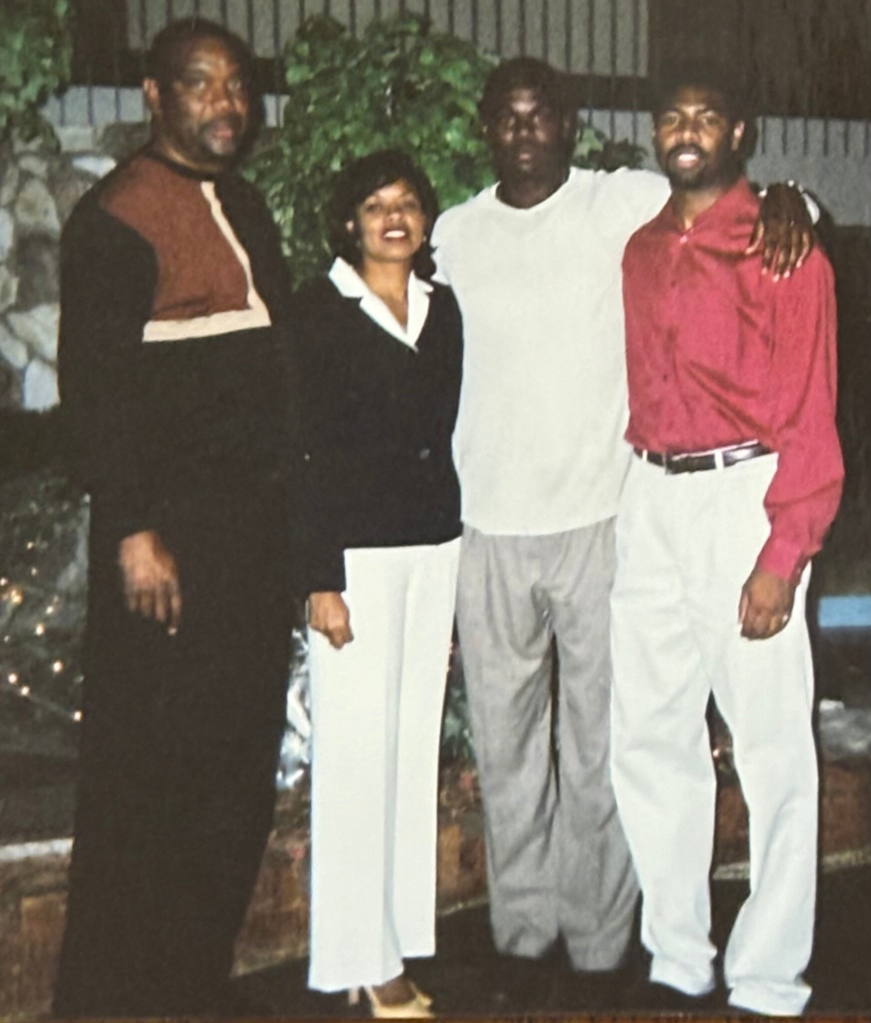 Four adults posing together outdoors at night, smiling in front of a hedge fence. Left to right: man in brown and black outfit, woman in black suit with white blouse, man in white shirt, man in red shirt with arm around his friend.