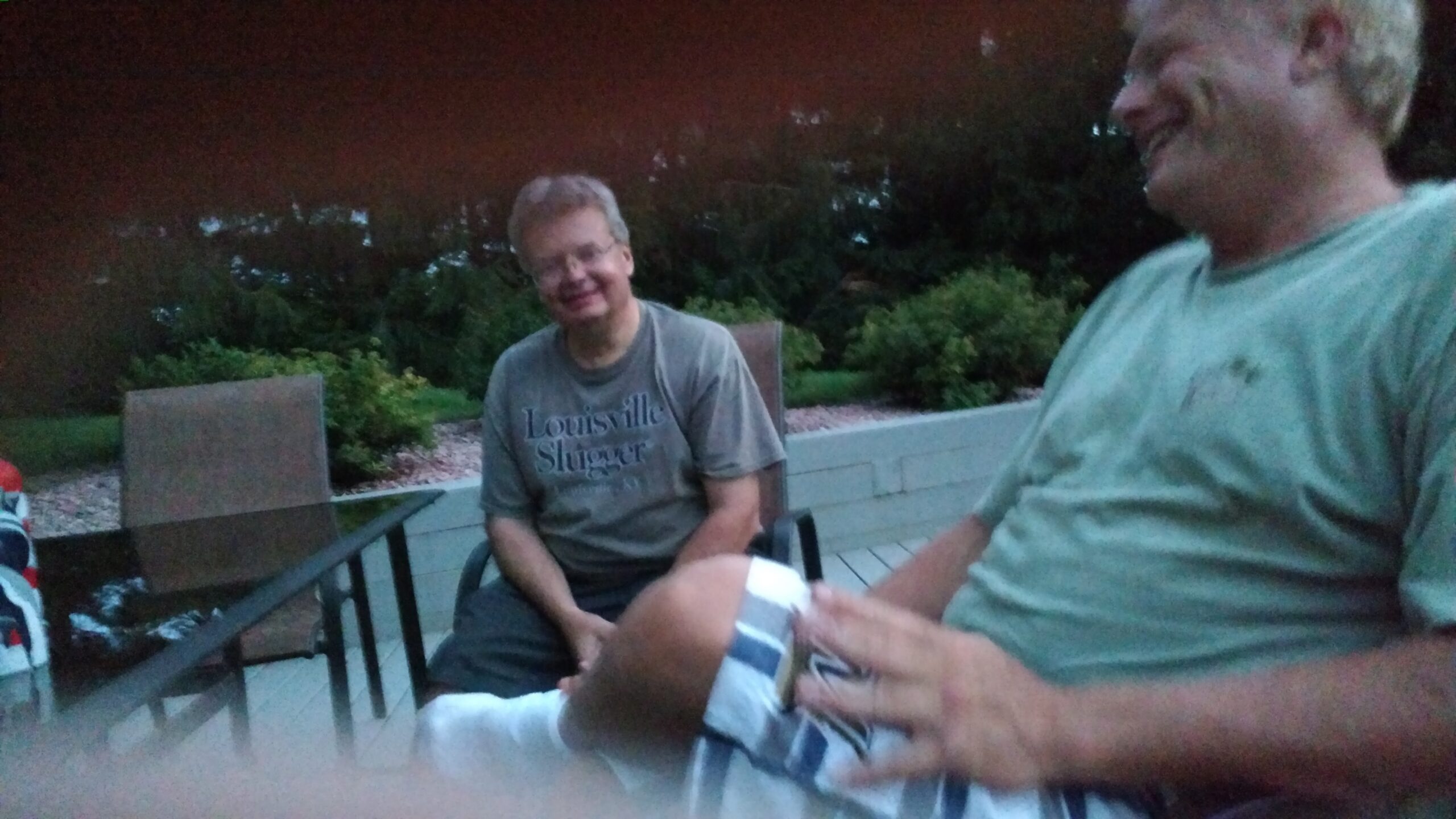 Two men sit on a backyard deck chatting at dusk; one in a Louisville Slugger shirt smiles while the other, in a green shirt, is partially in frame.