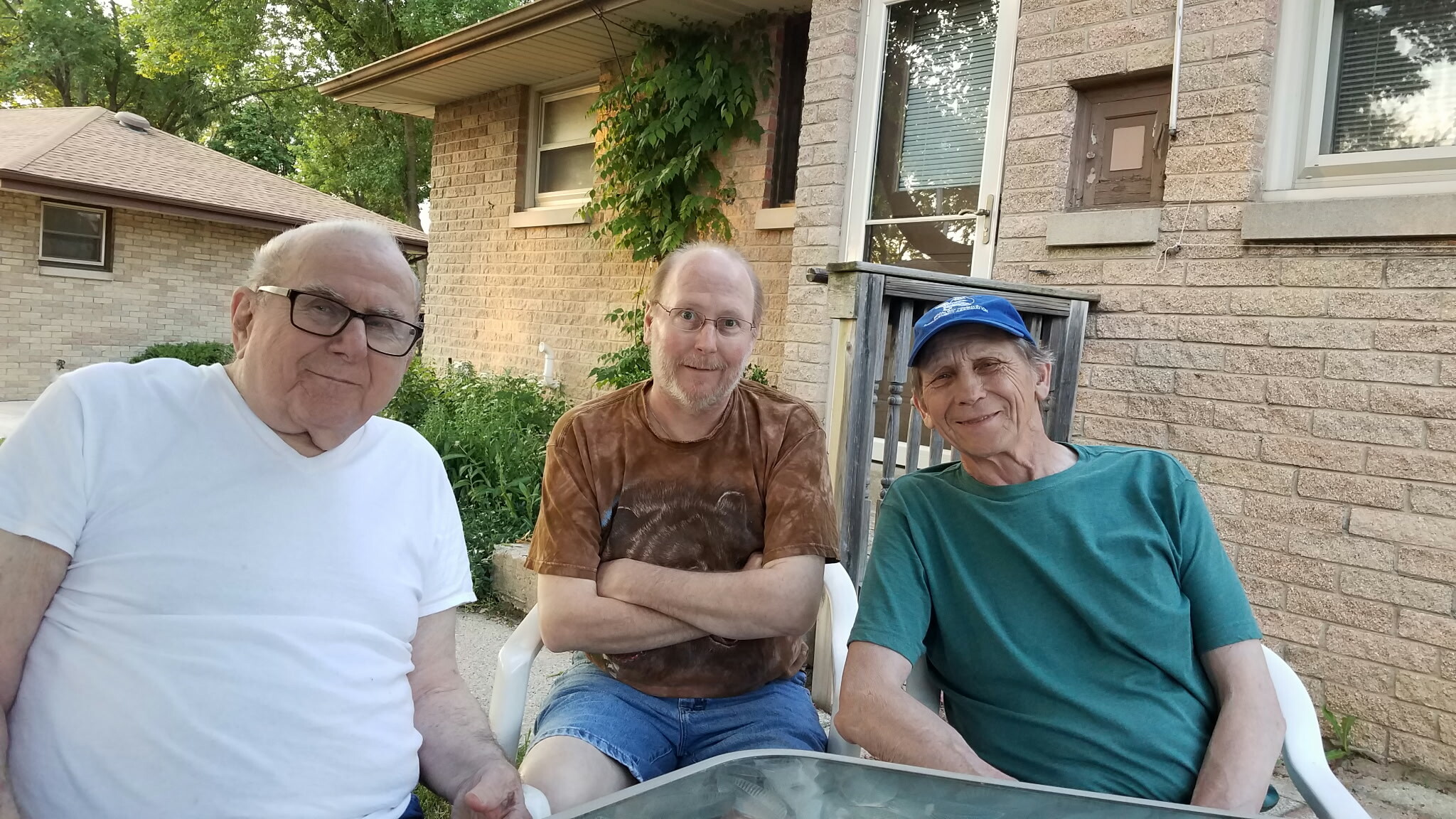 Three men sit together on white lawn chairs outside a brick house, smiling at the camera with trees and greenery in the background.