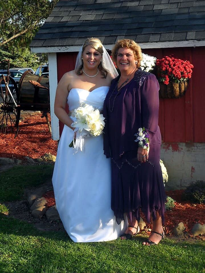 Bride in a white wedding gown holding a bouquet with a smiling woman in a purple dress beside her, outdoors by a red building with flowers