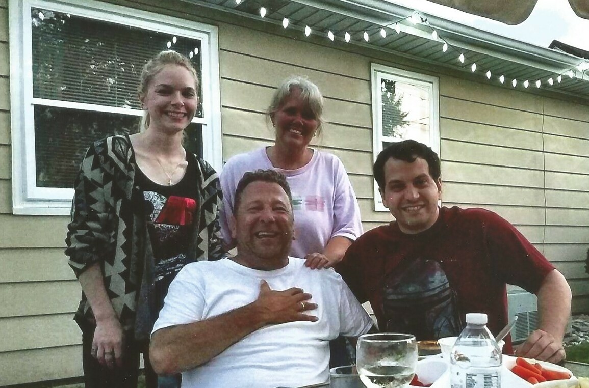 Four people posing together at an outdoor family gathering, smiling around a table with drinks on a sunny day by a house porch.