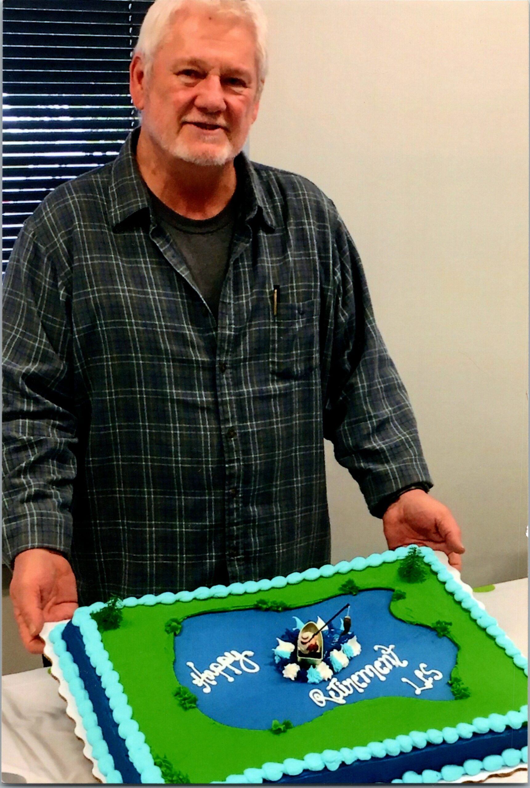 Older man with white hair and beard in a plaid shirt stands beside a large blue-and-green retirement cake with decorative frosting.