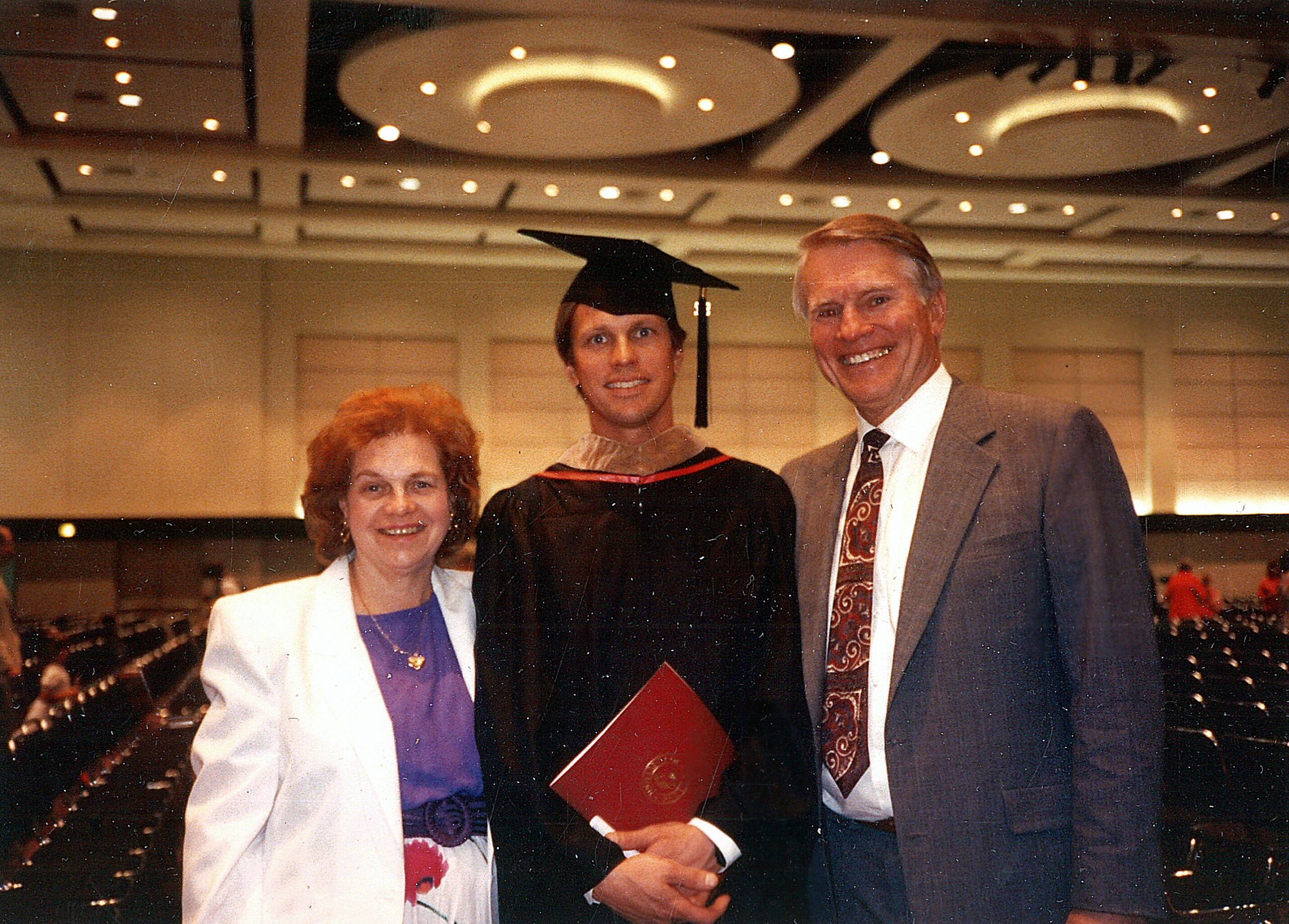 Graduate in cap and gown posing with his parents at a graduation ceremony.