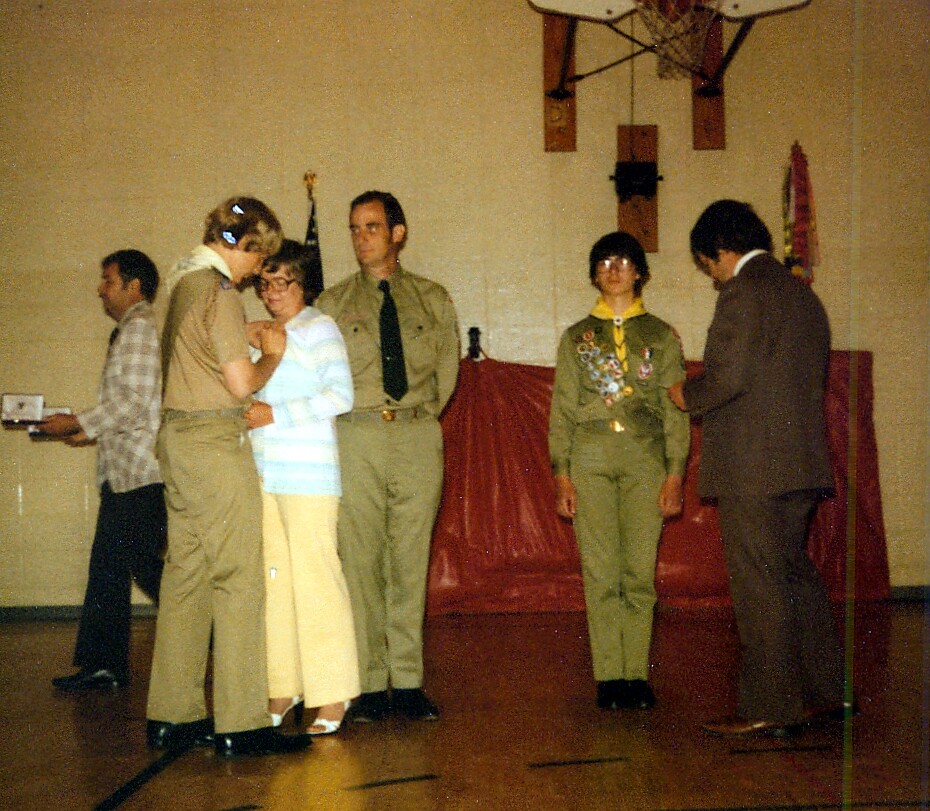 Group of people in a school gym on stage; a woman in light striped top receives a pin from a man in tan uniform while others watch.