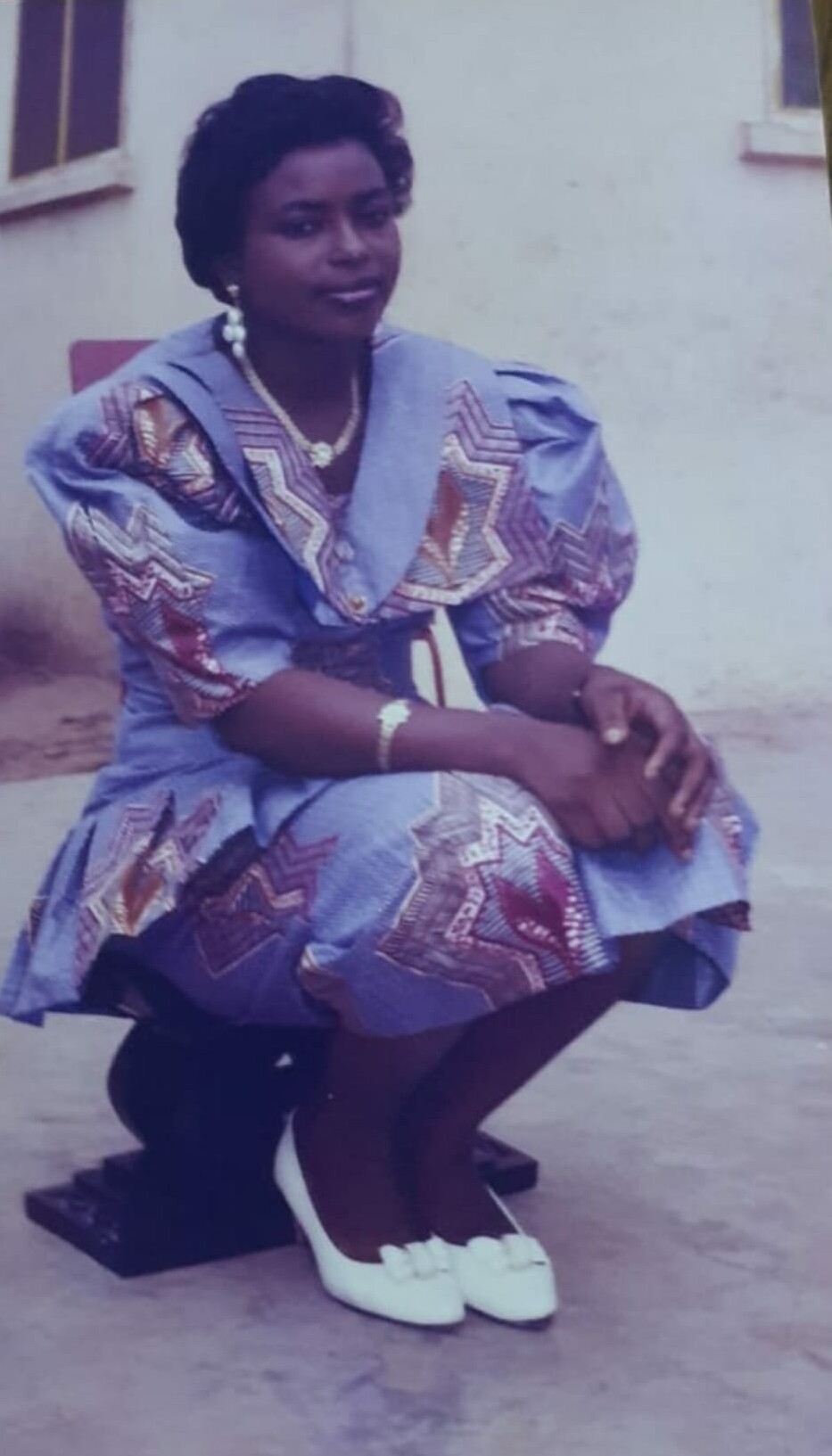 Woman in a blue patterned dress crouching outdoors on a concrete surface, wearing white heels, a gold necklace, earrings, and bracelet.