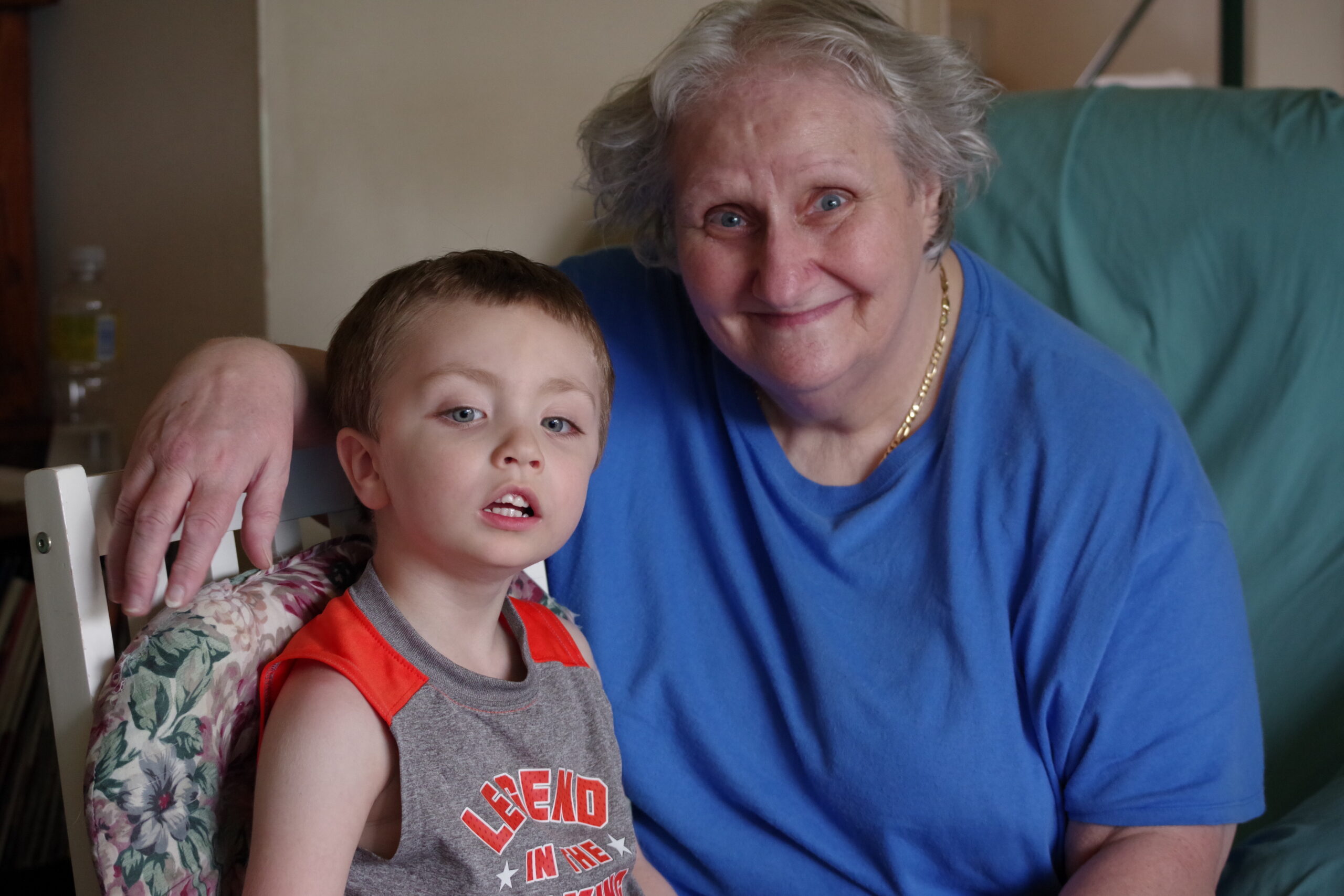 Grandmother and young boy sit closely together on a chair, smiling at the camera in a home setting.