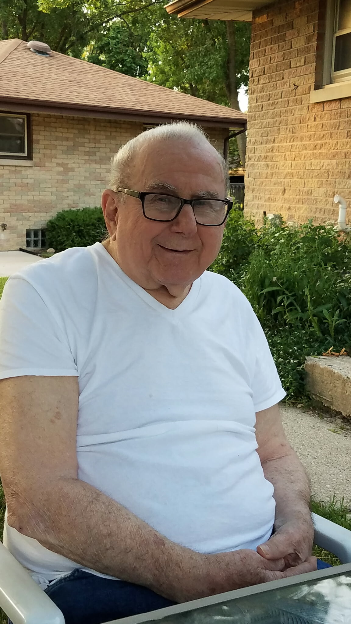Older man with white hair and glasses, wearing a white T‑shirt, sitting outdoors by a brick house.