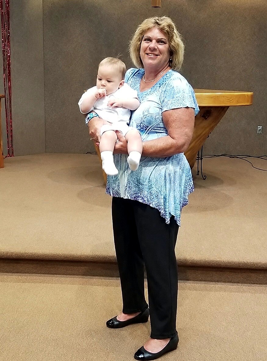 Older woman in a blue patterned blouse holds a small baby in white clothing, smiling at the camera in a stage setting.