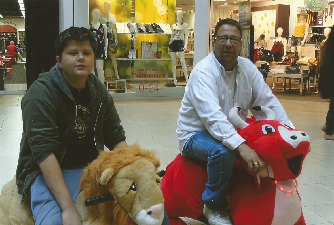 Two people riding large plush animal rides in a shopping mall: a boy on a tan lion and a man on a red dragon, storefronts in the background.