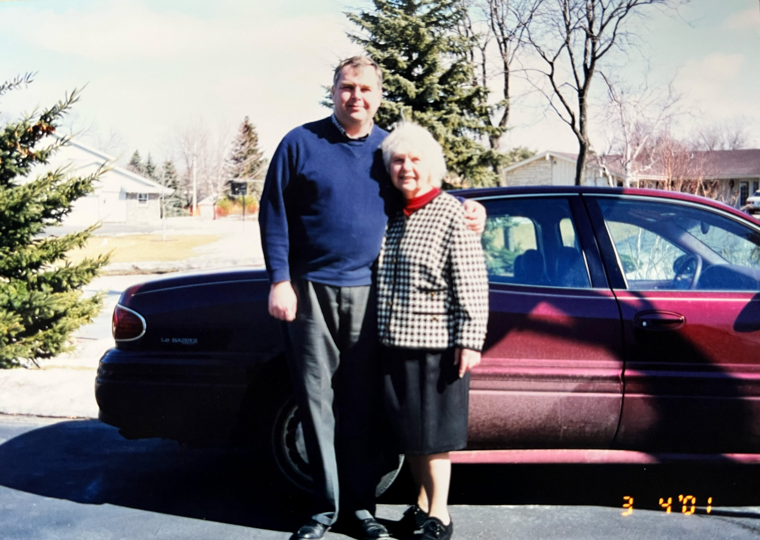 Two people standing together in a driveway beside a maroon car in a suburban neighborhood, smiling at the camera (man and older woman).