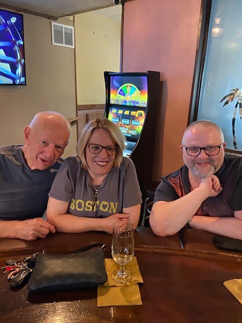 Three friends sit at a bar, smiling, with a glass of wine in front and a colorful slot machine behind them in the background.