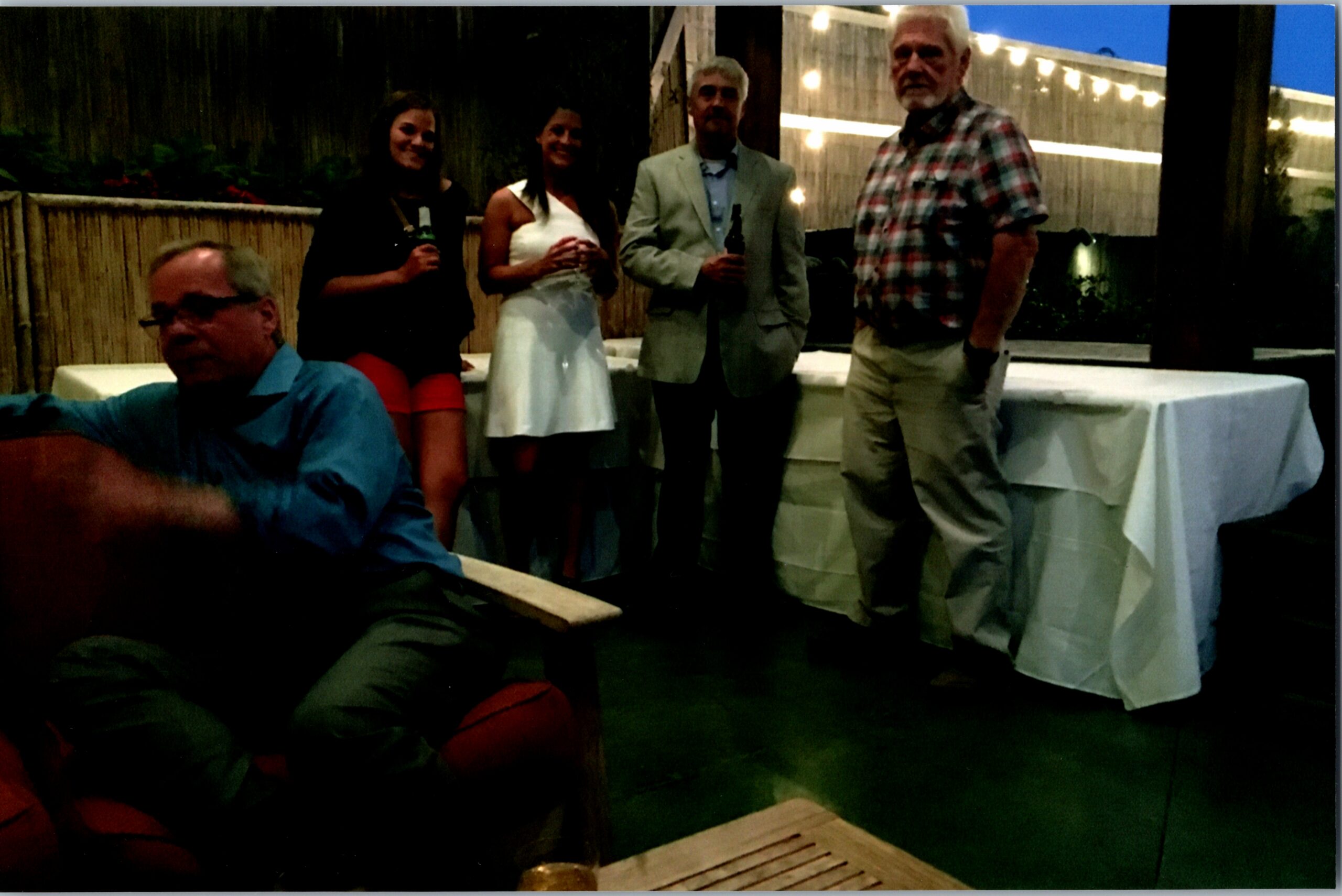 Five adults pose at an outdoor evening gathering, holding drinks near a table with string lights and a wooden fence in the background.