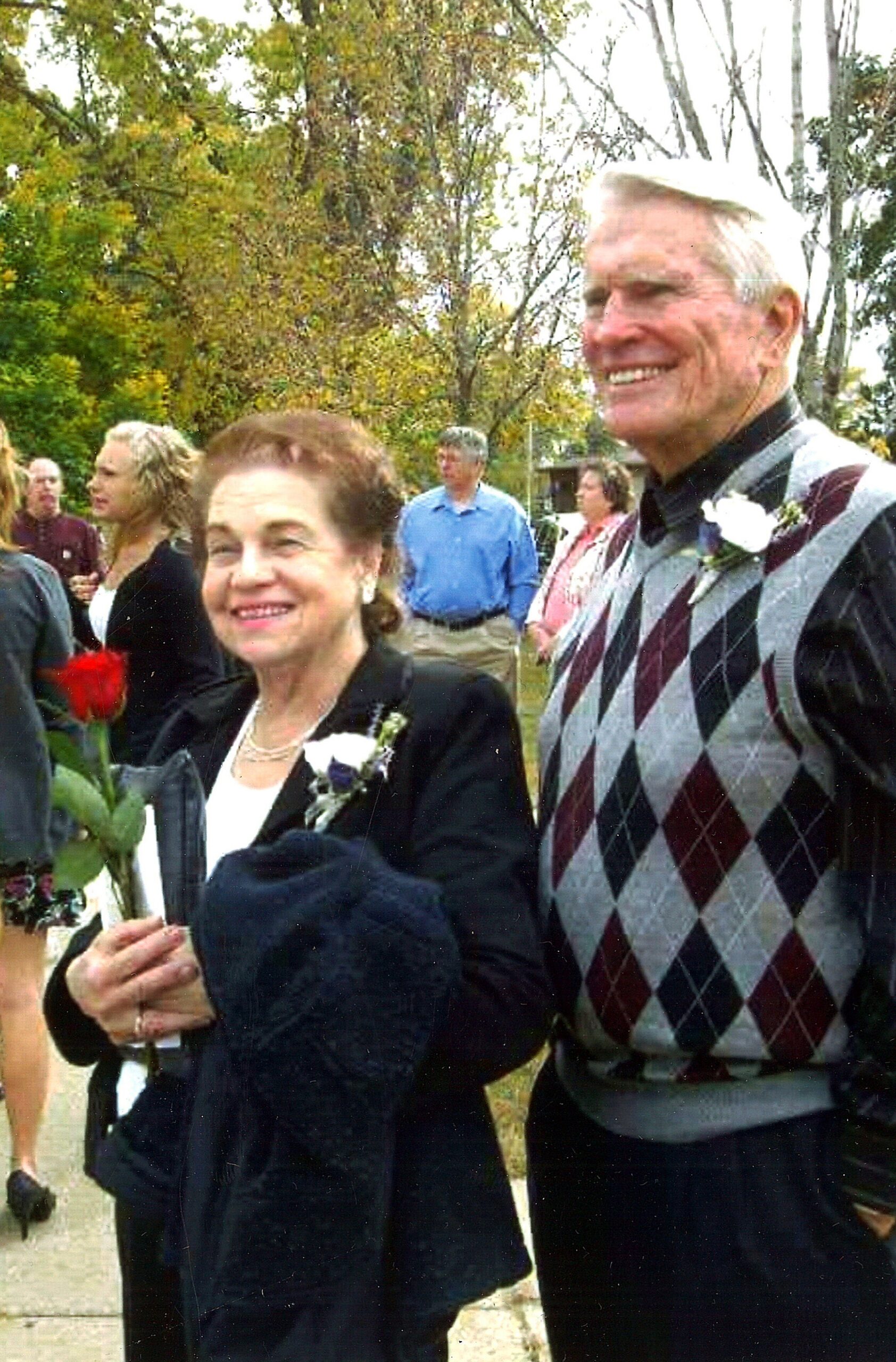 Smiling elderly couple at an outdoor gathering; woman holds a flower and corsage, man wears a maroon-gray argyle sweater vest in the foreground.