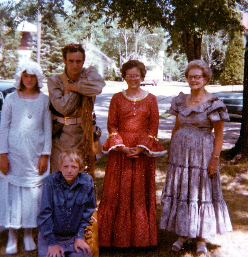 Five people stand outdoors in vintage costumes: a knight-like figure, a girl in a white nightgown, two women in long period dresses, and a boy in blue kneeling in front.