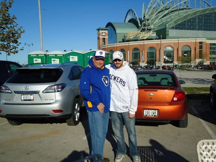 Two men pose together in a parking lot in front of a stadium with a green roof, one wearing a Brewers jersey and the other a blue jacket.