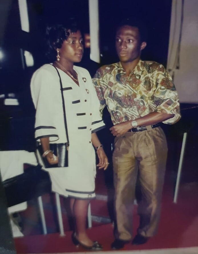 Two adults stand together at a social event: a woman in a light coat with black trim and pearl jewelry, carrying a black purse, and a man in a colorful patterned shirt and tan trousers.