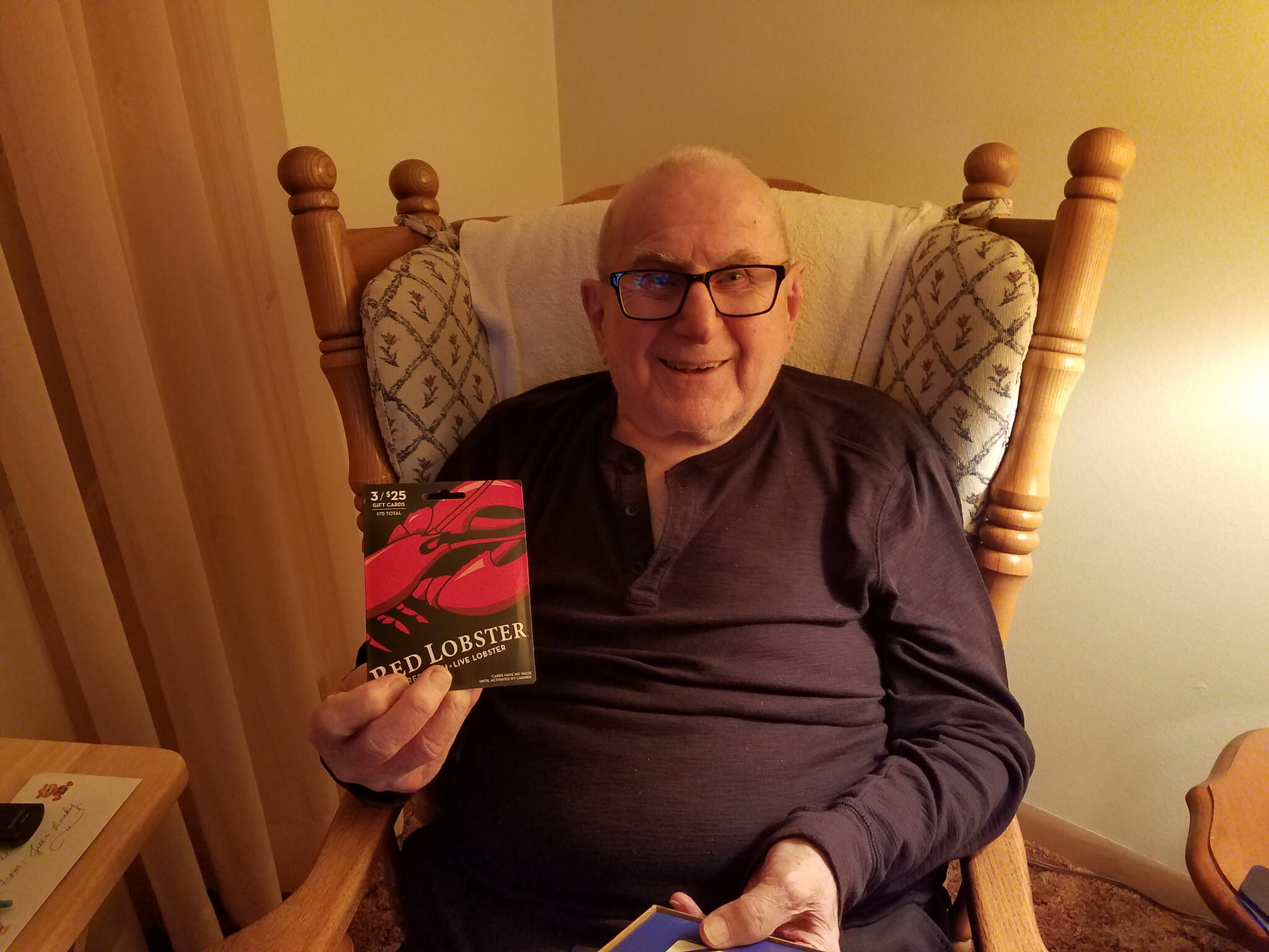 Smiling older man with glasses sits in a wooden armchair, holding a Red Lobster gift card toward the camera.